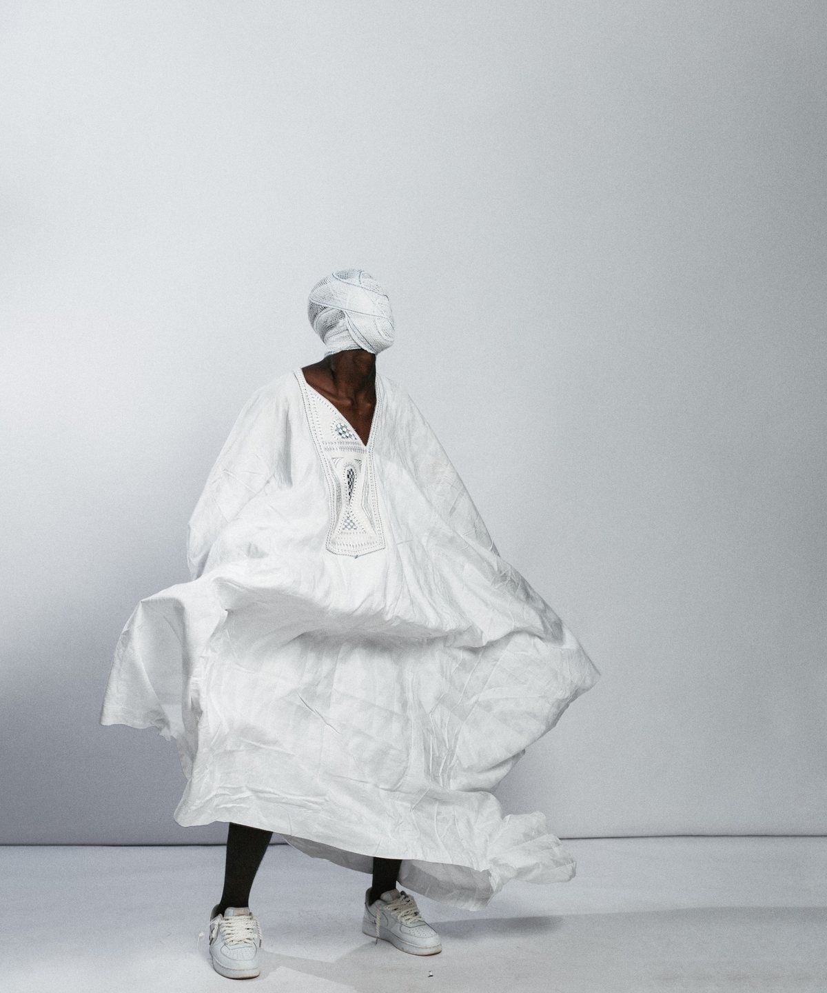Figure in a flowing white robe and headwrap, mid-turn against a pale studio wall