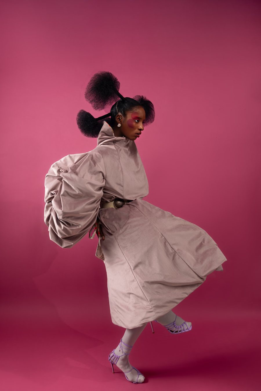 Model in a sculptural velvet coat kicking a heel up against a pink backdrop