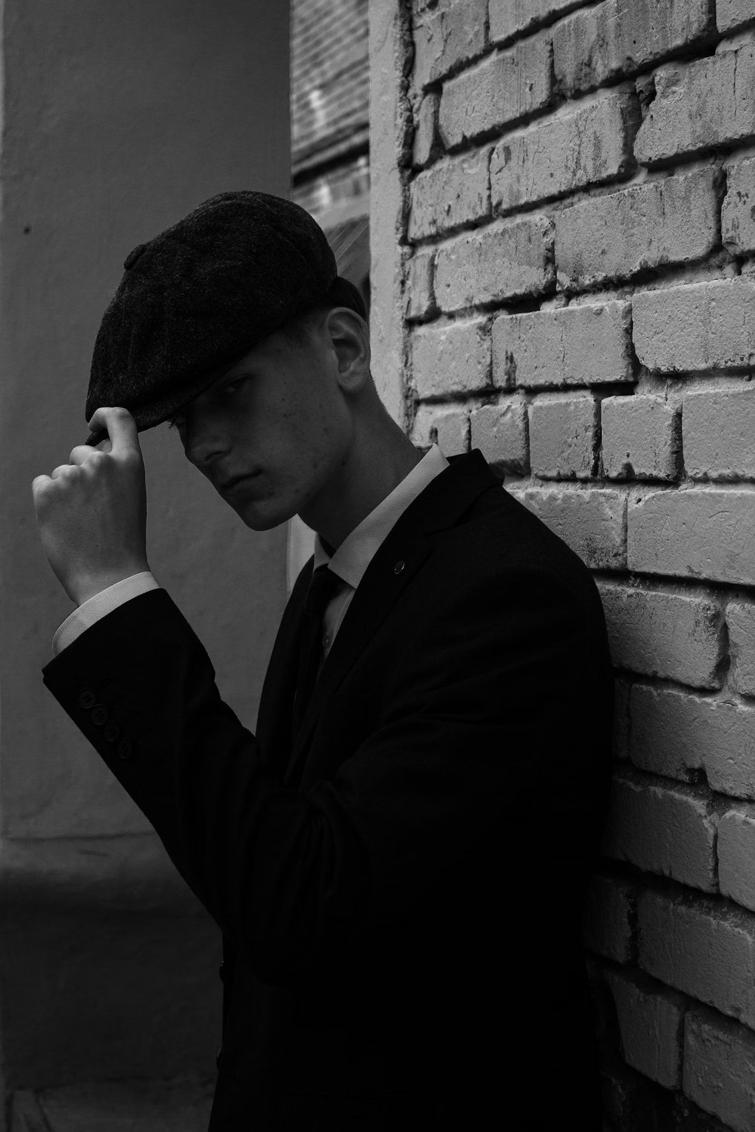 Man in a worsted suit and flat cap leaning against a painted brick wall