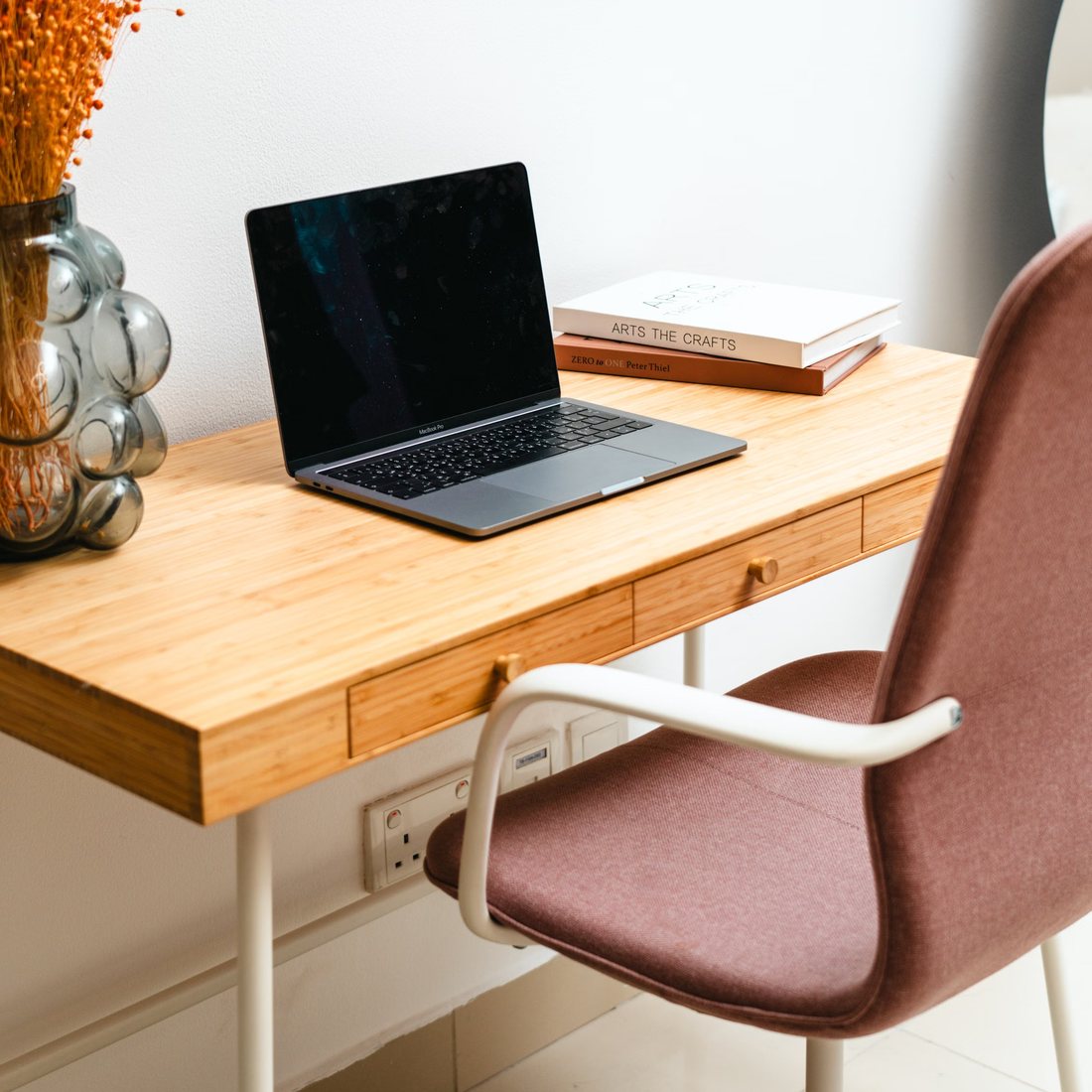 A bamboo desk with an open laptop, two books, and a soft rose chair