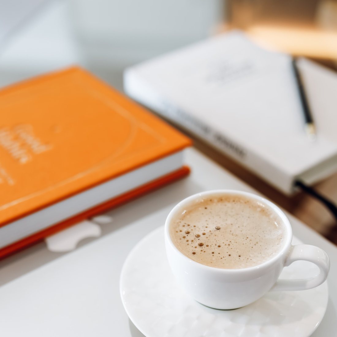 An orange notebook, a white book, and a cup of coffee on a light table