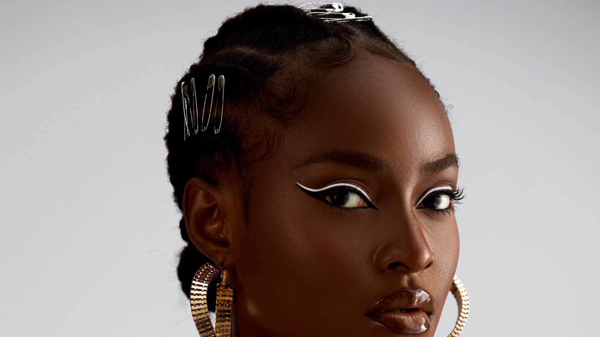 Studio portrait of a woman with graphic white eyeliner and gold earrings against a grey backdrop